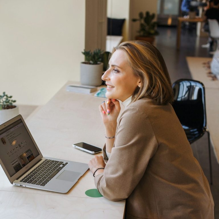 Frau sitzt an einem Tisch mit Laptop und Smartphone, blickt nachdenklich und lächelt dabei.