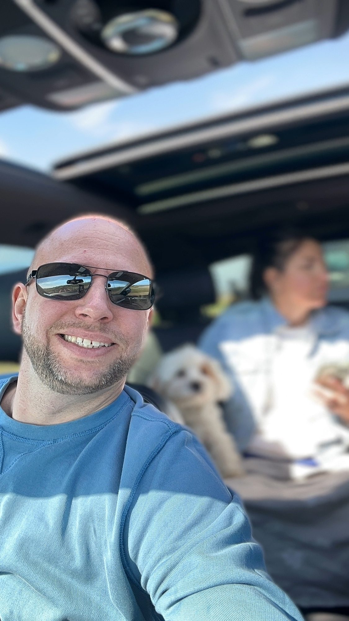 Mann mit Sonnenbrille lächelt im Auto, eine Frau und ein Hund sitzen im Hintergrund