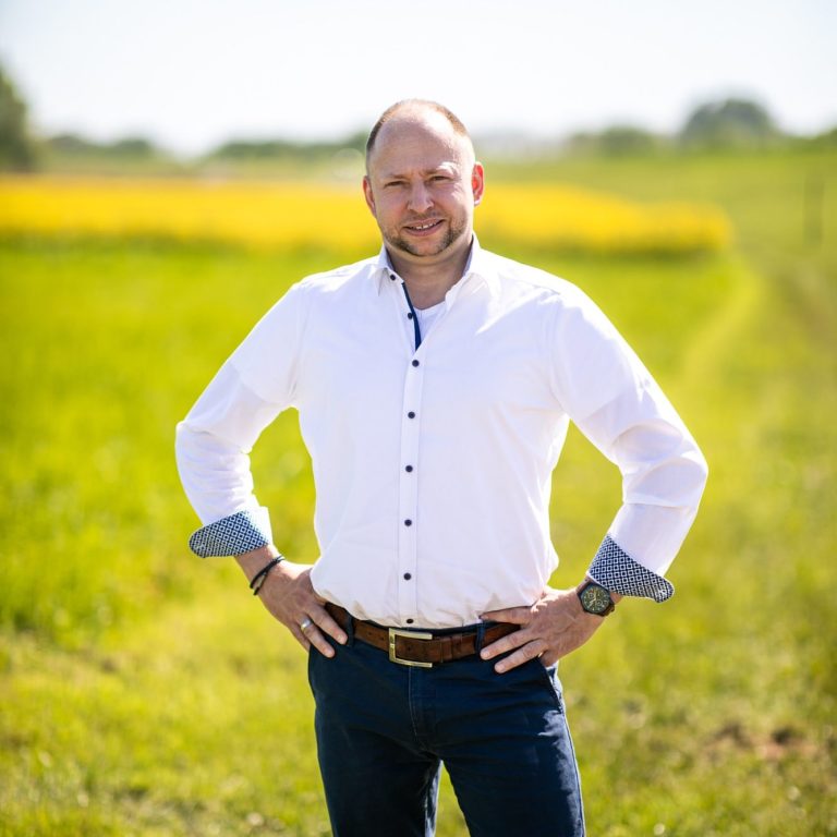 Mann in weißem Hemd und Jeans steht lächelnd auf einem Feld mit grüner Wiese und gelben Blumen.